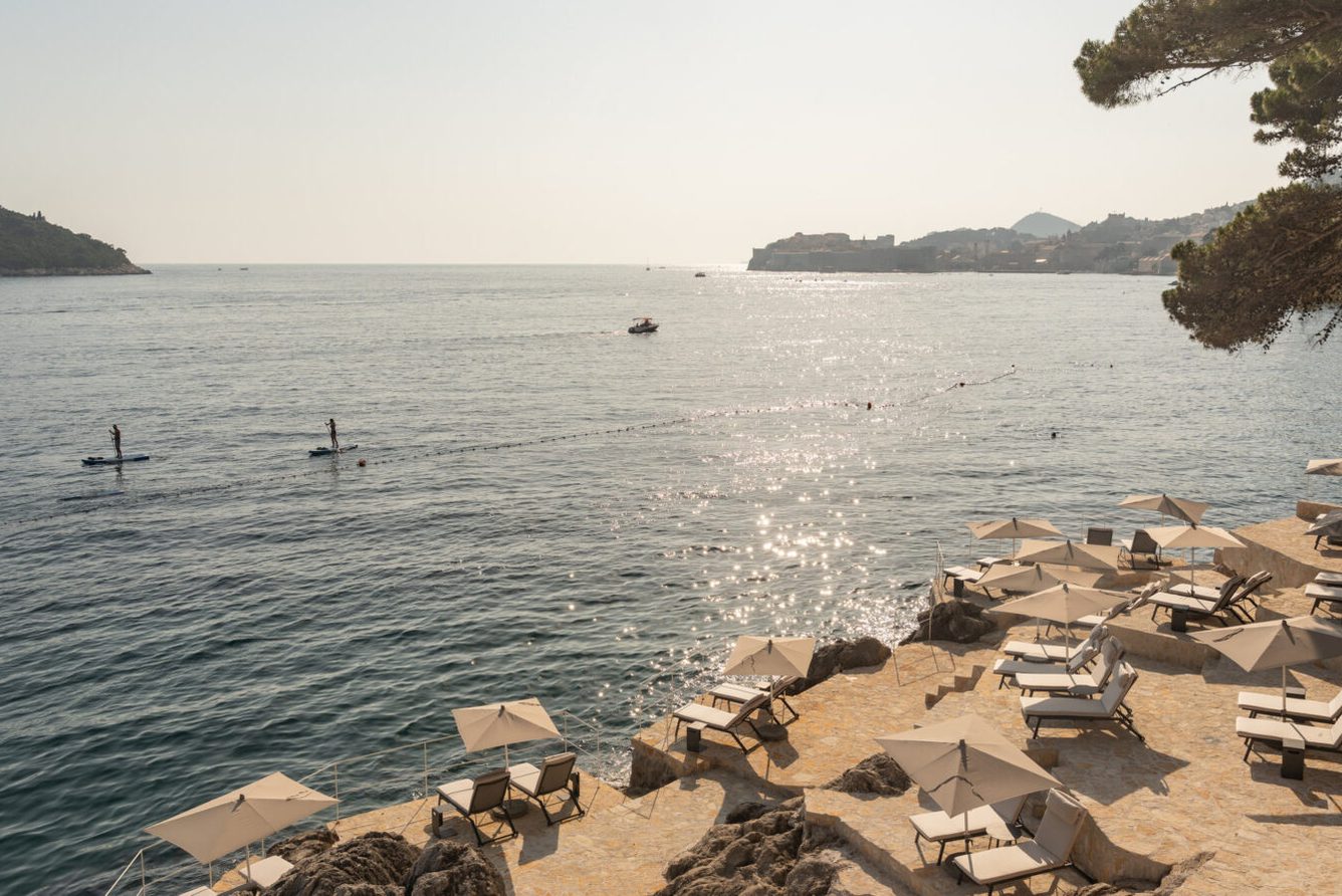 Villa Dubrovnik's beach terraces with sun loungers and parasols on stone platforms above the Adriatic, looking towards Dubrovnik Old Town