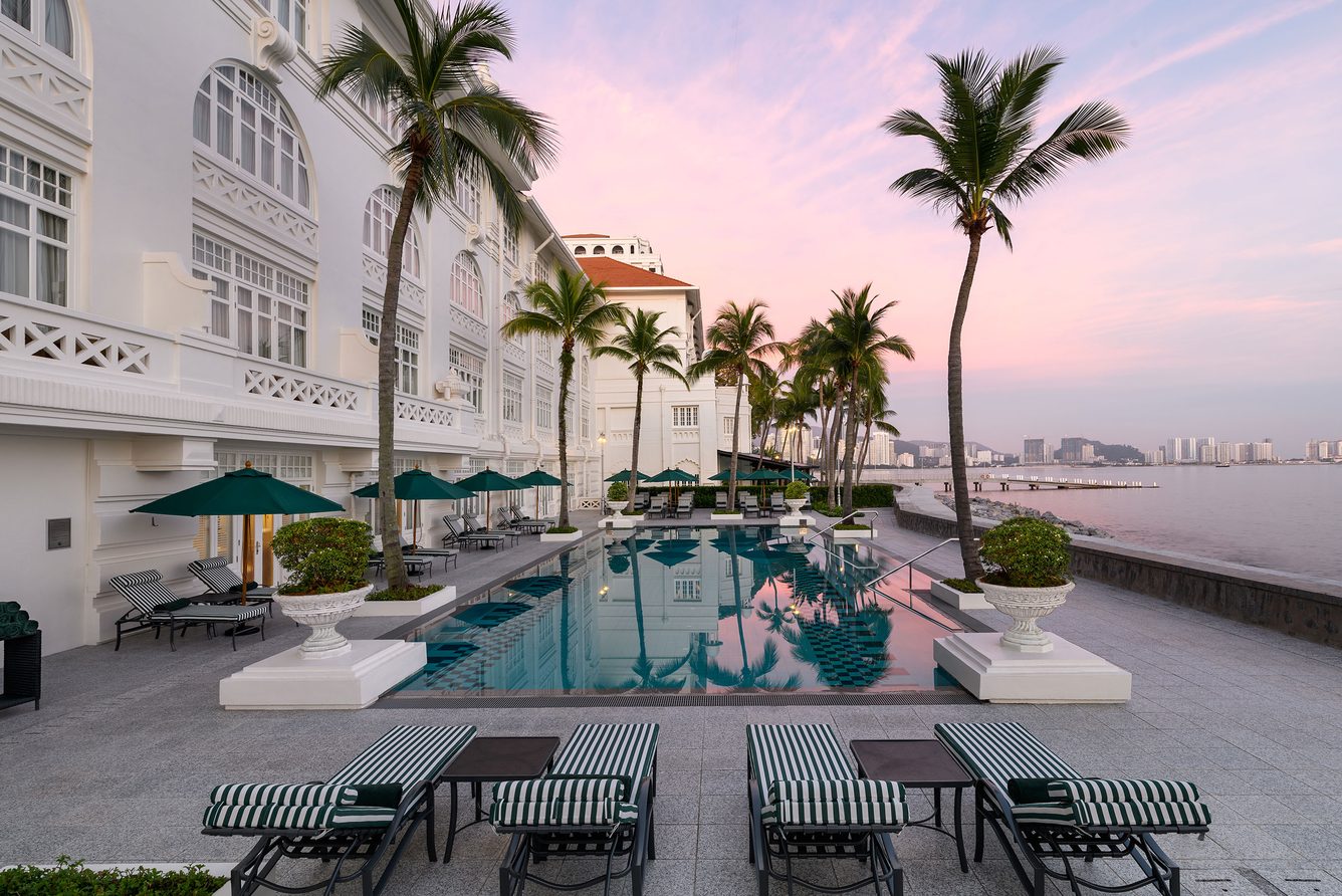 The Heritage Wing swimming pool at the Eastern & Oriental Hotel in George Town, with the hotel's white colonial façade, palm trees and the Penang Strait at dusk