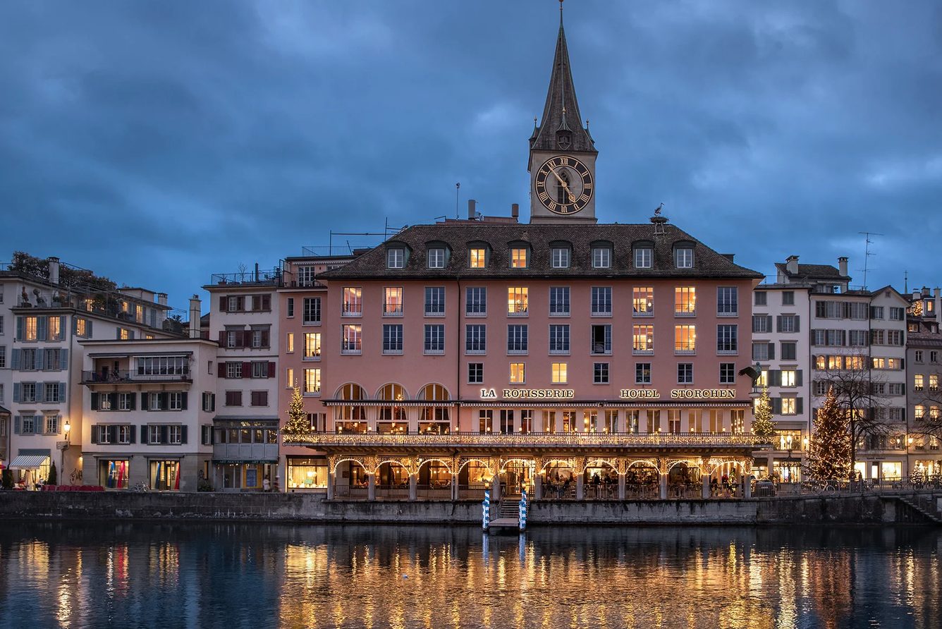 Storchen Zurich lit up at dusk across the Limmat in Zurich, below the tower of St Peter's church