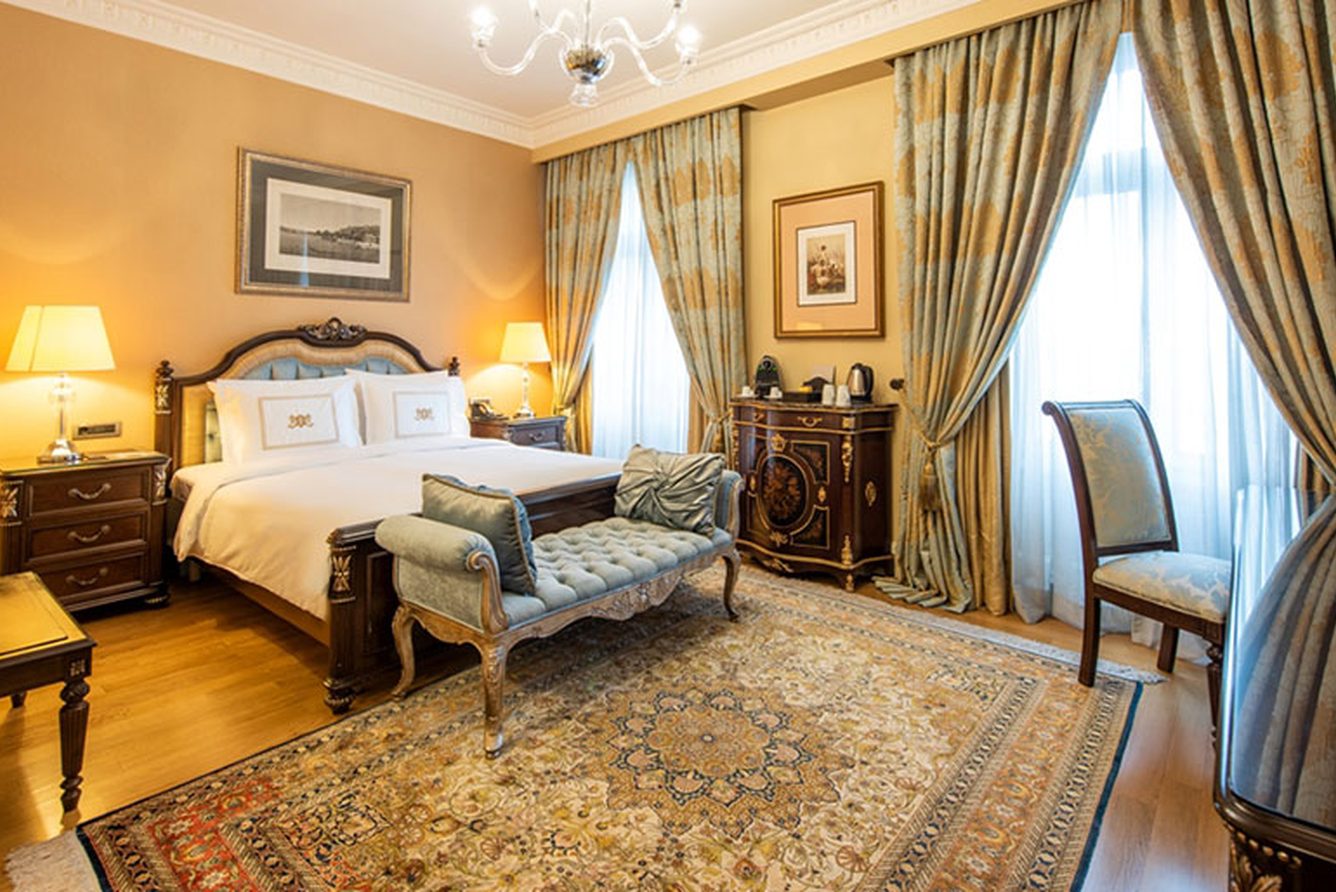 A suite bedroom at Pera Palace with a carved wooden bed, patterned rug and tall draped windows
