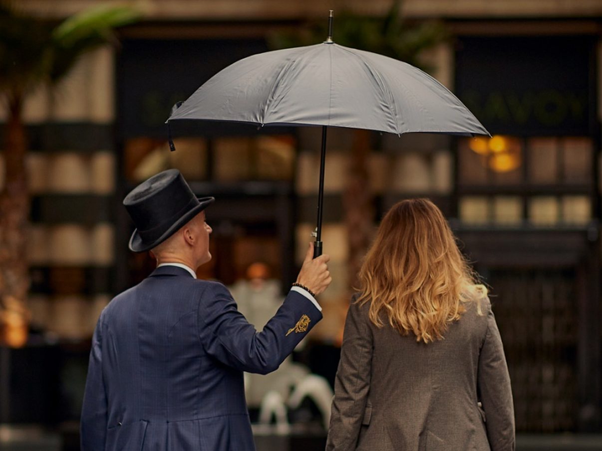 A Savoy doorman in a top hat holding an umbrella for guests outside the hotel on the Strand at night