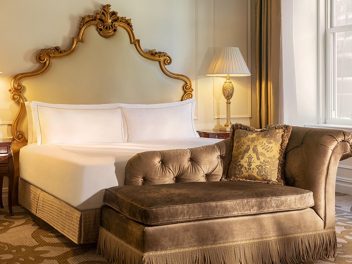 A suite bedroom at The Plaza with an ornate gold headboard, crisp white bedding and a chaise longue